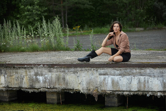 woman is sitting on a dock with one leg hanging over the edge. She wears a brown shirt and black shorts. area has tall grass and green plants. It is bright outside
