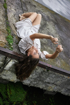 young woman is lying on a rocky surface beside the water. She is wearing a simple outfit and is holding her phone to take pictures of her surroundings during the day