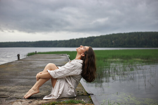 woman sits on a dock by a lake with grass in the water. She is barefoot and leans back with her eyes closed. Clouds fill the sky, indicating potential rain