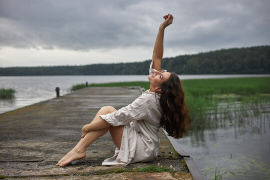 woman sits on a wooden dock by a lake. She wears a light shirt and holds her arm up. sky is cloudy and a green landscape is seen in the background during late afternoon