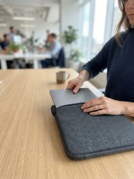 Woman Placing Laptop into Sleek Protective Sleeve at Bright Modern Office Desk with Blurred Colleagues Working in the Background Atmosphere
