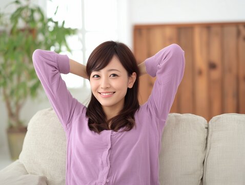ソファでリラックスしながら頭の後ろで手を組み笑顔を見せる紫色の服を着た若い女性