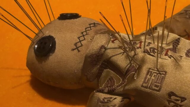Voodoo doll made of fabric with needles stuck in it. Traditional mystical object for magic ritual. Dark occult theme and spiritual symbol on orange background.