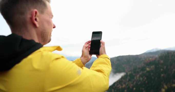 Standing on a breathtaking mountain peak, a man in a bright yellow jacket takes stunning photos with his smartphone. Wispy clouds float below him as he embraces the serene view.
