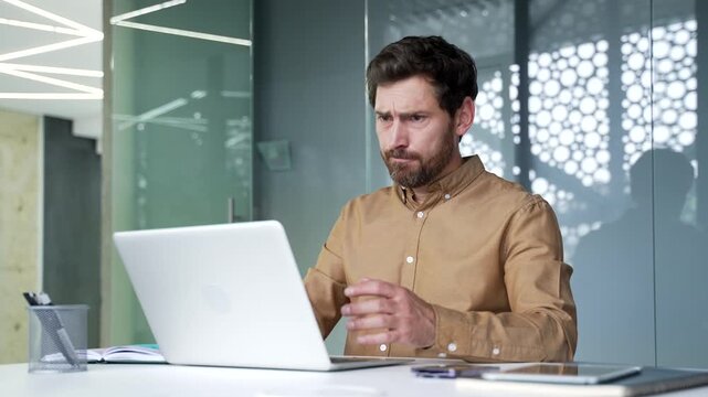 Frustrated mature businessman struggling with a computer problem on his laptop in a modern office. Angry professional reacts to a technical issue while working at his desk in a corporate workplace.