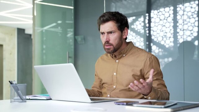 Angry mature businessman arguing during an online video meeting on a laptop at his desk in a modern office. Frustrated manager expressing dissatisfaction while confronting a colleague remotely.