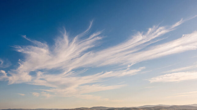 Graceful cirrus clouds streak across a soft blue sky above a distant, hazy mountain landscape in calm weather.