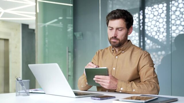 Focused mature businessman taking notes in a notebook while attending an online business seminar in office. Professional employee listening carefully to a virtual presentation and learning new skills.