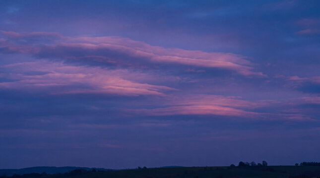 Overcast, dramatic twilight sky with swirling purple and pink clouds above rolling green hills landscape.