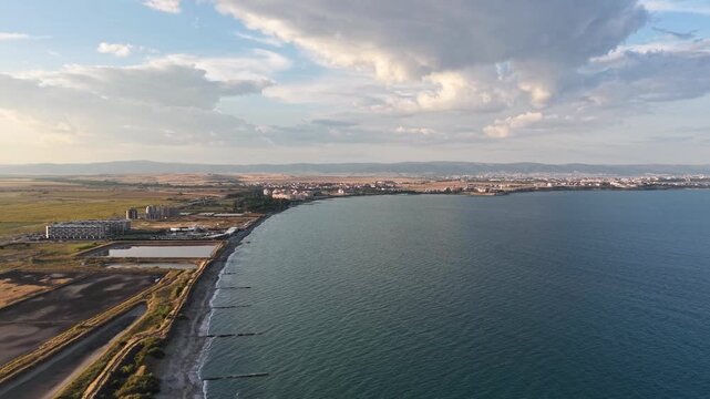 Wide-angle aerial drone shot revealing the picturesque coastline of Pomorie, Bulgaria. The clip features the calm blue Black Sea, sandy shoreline with breakwaters, saline evaporation pans, and the dis