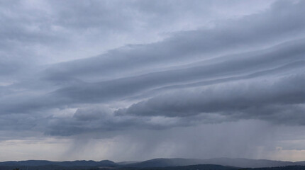 Naklejka premium Expansive gray sky with striking, layered undulatus clouds and heavy rain falling over distant hills.