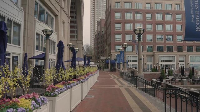 A brick-paved pedestrian promenade lined with colorful spring flowers, outdoor dining areas, and closed blue umbrellas in an upscale urban waterfront district.