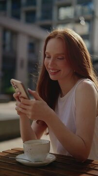 Beautiful redhead woman happily texting on mobile phone enjoying cappuccino at sidewalk cafe on sunny day. Vertical portrait of smiling female communicating on social media, dating app outdoors.