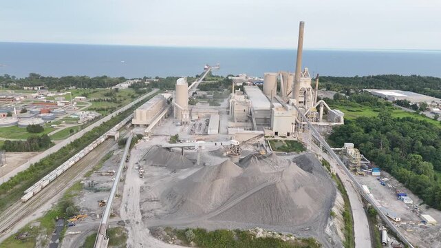 An aerial drone shot flies forward over large gravel mounds, heading toward the processing infrastructure, silos, and smokestack of the CRH Ash Grove cement plant
