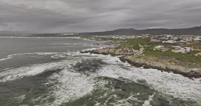 Gansbaai South Africa Aerial v2 low flyover around De Kelders homes above a rocky coastline with waves, mountains and overcast skies - Shot with Mavic 3 Pro Cine - Jan 7th 2024