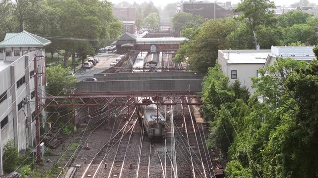 Commuter train approaches historic railway station in New Jersey, overhead catenary wires cross tracks as platform sits between old brick buildings and dense green trees. Aerial drone wide shot