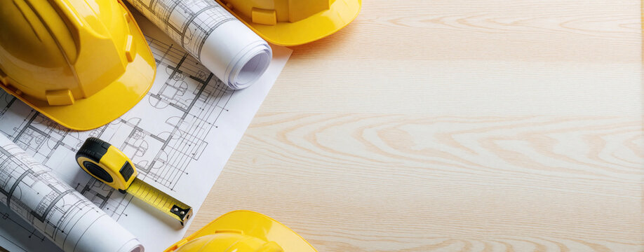 Construction safety equipment flat lay on wooden table. Yellow hard hats, rolled blueprints lie near architect building plans, tape measure. Pro tools for engineer, contractor builder during site