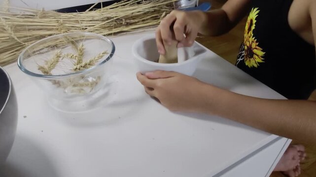 A high-angle side shot shows a student holding a white ceramic mortar and grinding wheat grains with a wooden pestle during a hands-on 3rd grade Waldorf school practical farming activity on a white de