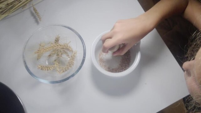 An overhead close-up shot shows a student using a wooden pestle to grind wheat kernels into coarse flour inside a ceramic mortar beside a glass bowl during a hands-on Waldorf 3rd grade farming lesson.
