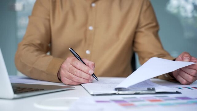 Close-up of a businessman hand in a shirt signing business documents at a desk in a modern office. Professional executive approving an important contract and completing official paperwork.