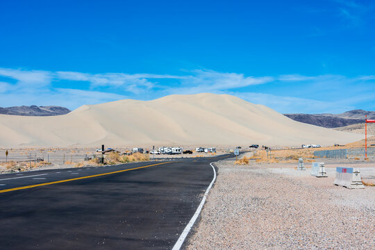 View of the Sand Mountain dunes in the Sand Springs Range with an asphalt road leading towards parked recreational vehicles under a clear blue sky in Churchill County, Nevada, United States.