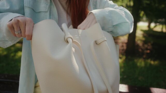 Cropped shot of woman with fresh manicure opening stylish beige tote bag and rummaging through contents, searching for item relaxing outdoors in beautiful sunny park. Shooting in slow motion.