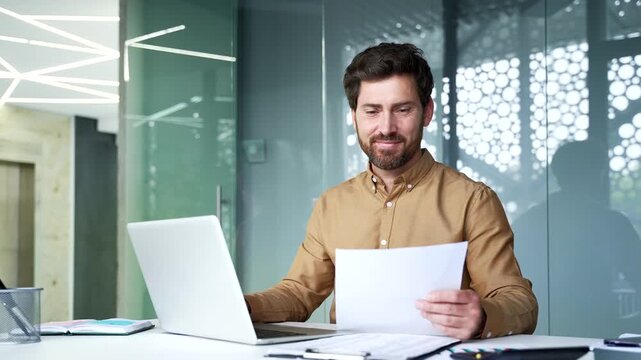 Happy mature businessman looking at a laptop while holding a financial report at his desk in a modern office. Smiling financier pleased with strong business performance and excellent financial results
