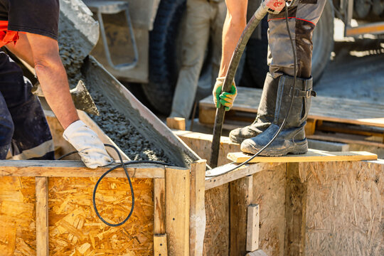 Workers are using a hose to pour concrete into a wooden form. One worker stands on the form while another supports the hose.