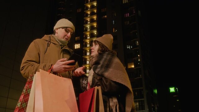 Caucasian young couple holding shopping bags and looking at smartphone screen on city street at night, low angle shot. Footage is perfect for holiday shopping or mobile banking promotions