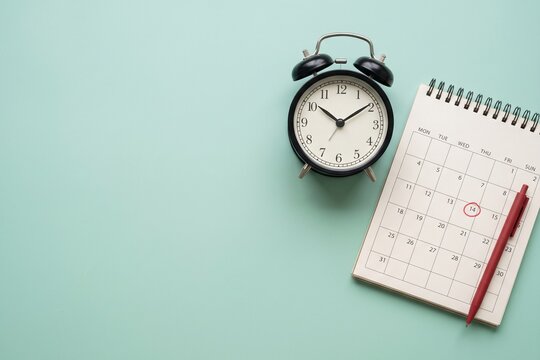 Alarm clock with open spiral notebook calendar and red pen on light green background top view copy space