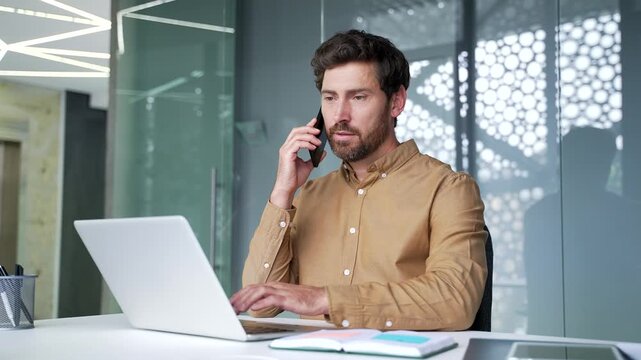 Mature businessman discussing work on a smartphone while reviewing information on a laptop at his desk in a modern office. Focused professional managing daily business tasks and communication.