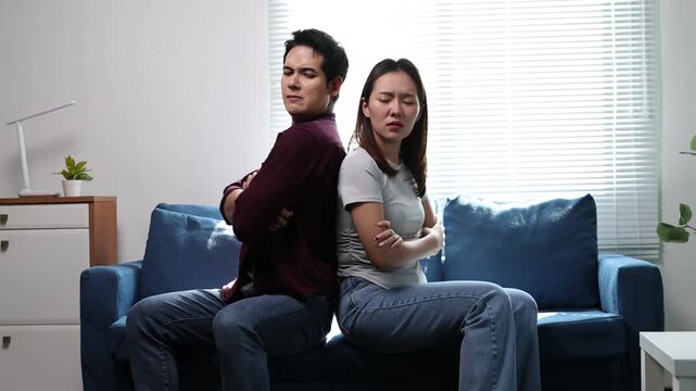 An Asian couple sits back to back on a living room couch, experiencing relationship tensions after an argument.