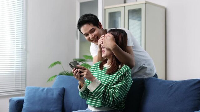 A boyfriend sneaks into his girlfriend's house, blindfolding her, to surprise her while she is playing on her phone and sitting on a blue sofa in the living room.