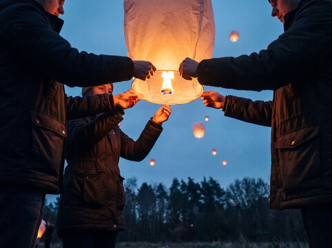 Group of three people releasing glowing sky lantern at dusk with other lanterns floating in dark blue sky