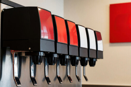 Row of soda fountain dispensers with red and white labels on a metal wall. A red square artwork hangs on beige wall. Ready drinks for customers.