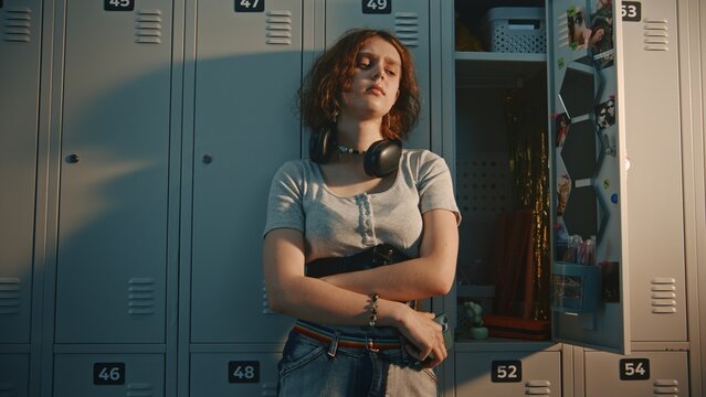 Teenage Schoolgirl With Headphones at Neck Standing in High School Locker Room. Girl Holds Smartphone, Staring at Screen With Upset Facial Expression While Reading Text Message or Social Media Post.