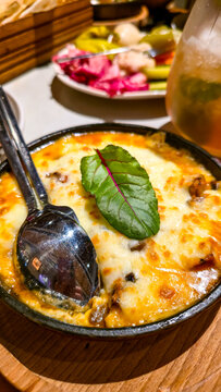 Vertical close-up shot of hot traditional baked cheese dish with golden melted crust garnished with fresh green leaf, served in a red cast iron skillet on a wooden board.