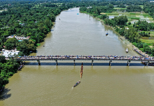 Aerial view of a traditional boat race on the river beneath a bridge crowded with spectators, surrounded by lush green fields and trees in Sirajganj, Rajshahi Division, Bangladesh.