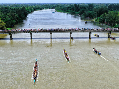 Aerial view of a traditional boat race with long wooden boats filled with rowers on the river, passing under a bridge crowded with spectators in Sirajganj, Rajshahi Division, Bangladesh.