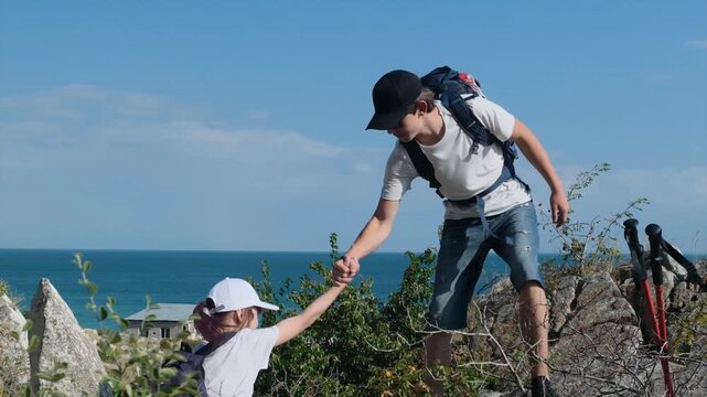 Teenage boy helps a girl climb a rocky mountain during a hike, pulling her up by the hand. Helping each other, hiking together, summer hiking and coastal mountain walks