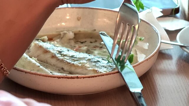 Hand with fork adjusts fresh herb garnish above fish fillet in creamy sauce inside shallow ceramic bowl on wooden table, showing careful final plating movement with warm daylight and rustic dining