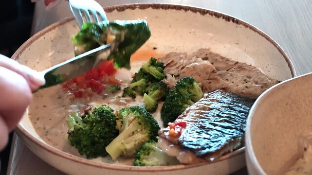 Hand with fork adjusts fresh herb garnish above fish fillet in creamy sauce inside shallow ceramic bowl on wooden table, showing careful final plating movement with warm daylight and rustic dining