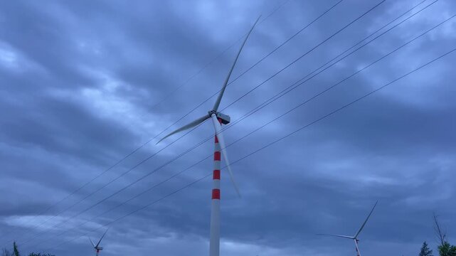 Handheld medium wide shot of wind turbines rotating at dusk with power lines under stormy sky in Quy Nhon Vietnam