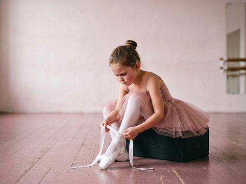 Young ballerina sitting on studio block tying ribbons of her pointe shoes