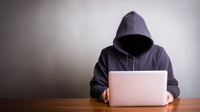 Hooded person using laptop in dark room, representing cyber crime, hacking, and online anonymity