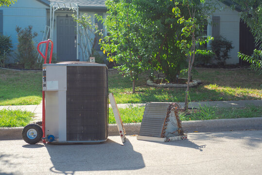 Retired air conditioner unit rests on red hand truck beside detached evaporator coil. Weathered metal fins, copper bends, and sunlit asphalt display residential service themes, Coppell