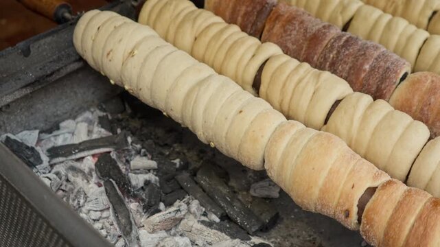 Traditional trdelnik chimney cake bakes on rotating wooden rolls over hot coals. Raw dough turns golden near glowing charcoal at a street food market. Fresh European sweet pastry cooks outdoors.