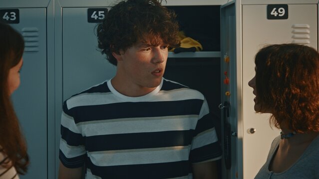 In Hallway Angry Teenage Girl Aggressively Grabs Male Classmate By Collar Of T Shirt. Boy Looks Shocked While Girls Corner Against Metal Lockers. Physical Abuse, Teenage Rage, And High School Assault.