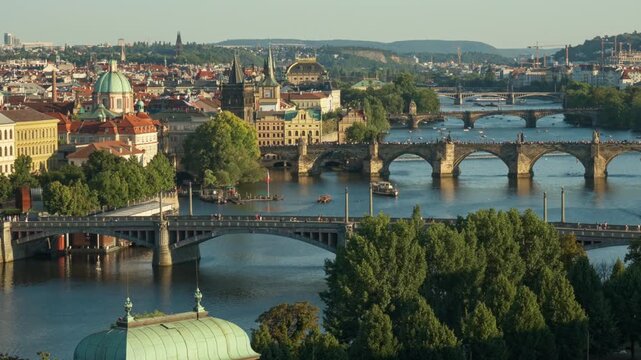 Panoramic view shows Vltava river and historic stone bridges in Prague city. Urban architecture and green trees line the embankment. Popular European travel destination in Czech Republic.