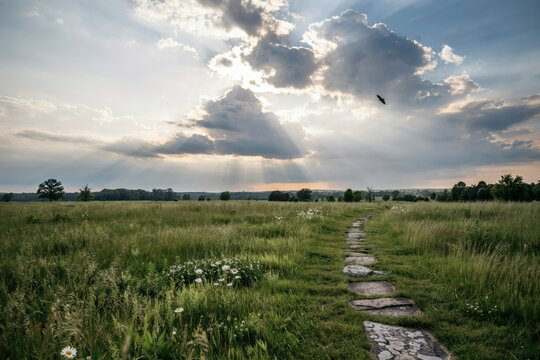 A winding stone path through a lush green grassy field under a dramatic cloudy sky with sun rays breaking through clouds in the late afternoon